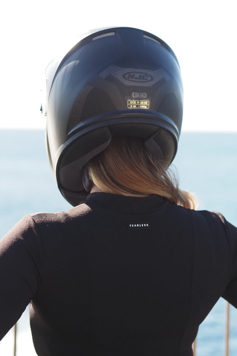 Wearing the Fearless au Cropped Rashie, a person in a black motorcycle helmet stands with their back to the camera, gazing out at the ocean.