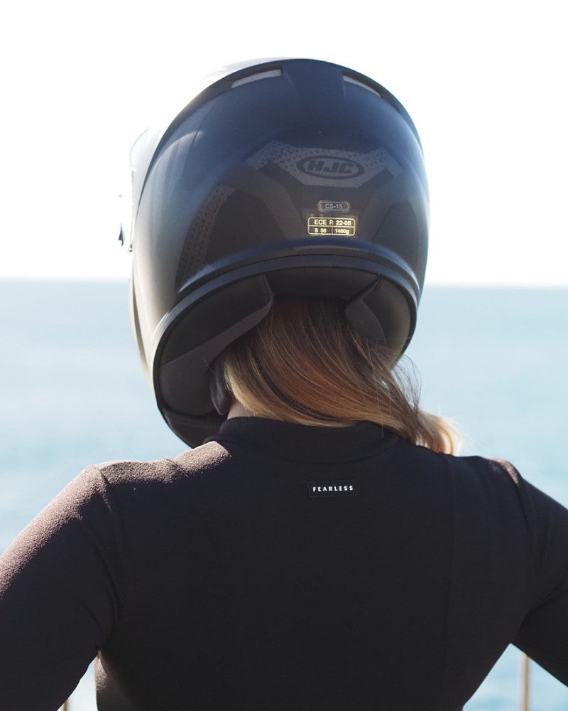 A person with long hair wearing a black motorcycle helmet and a black shirt stands facing the ocean, with their back to the camera on a sunny day.