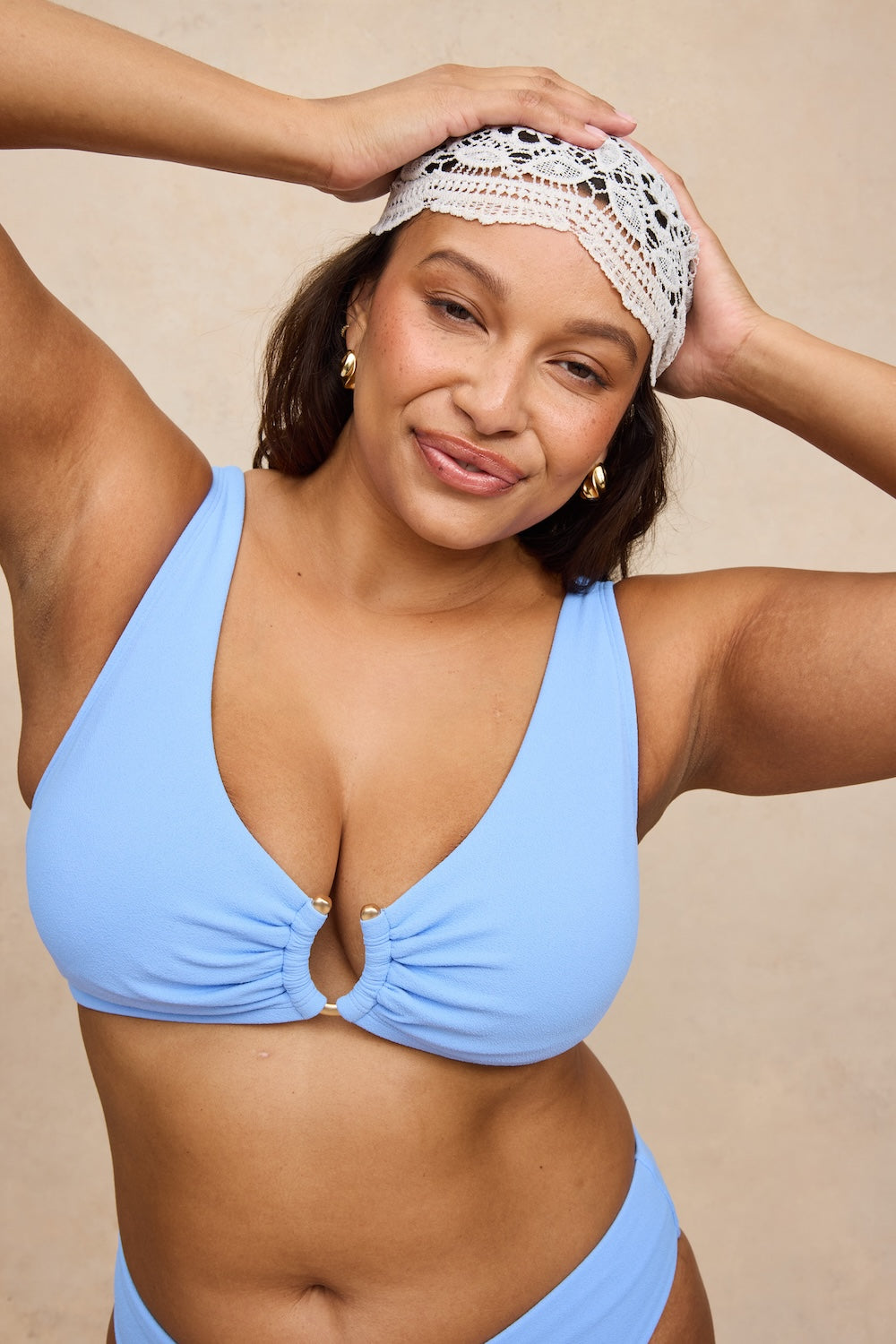 A woman wearing a light blue bikini top and matching bottoms, with a white lace scarf, posing against a neutral beige background.