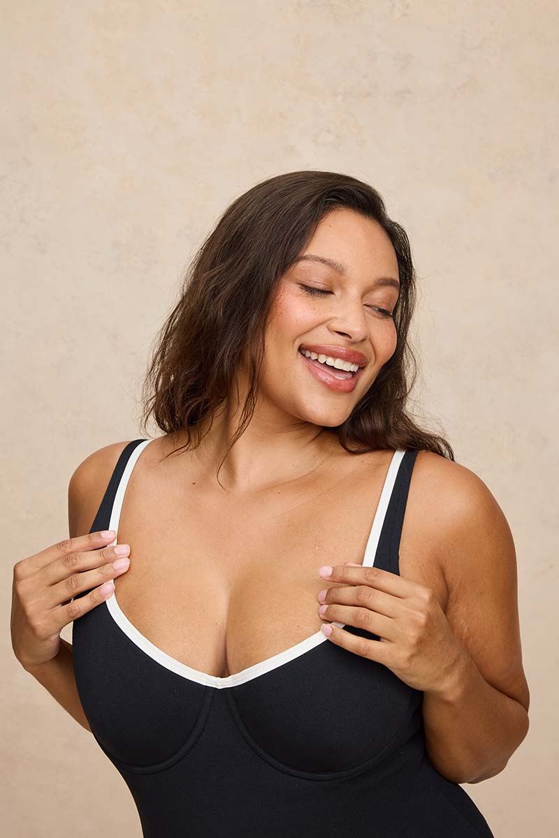 A woman smiling and posing in a black scoop-neck bralette with white trim, set against a textured beige background.
