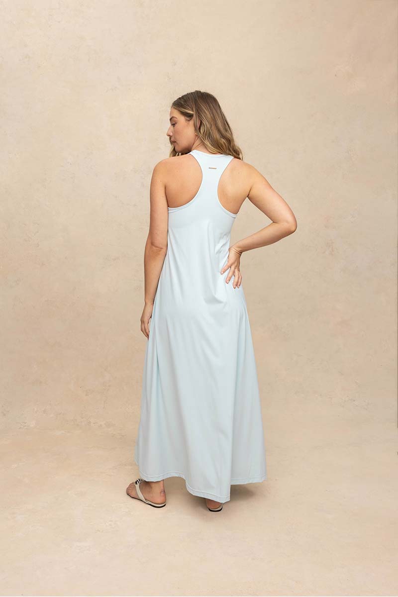 A woman modeling a light blue sleeveless maxi dress from the back, standing against a textured beige studio backdrop.