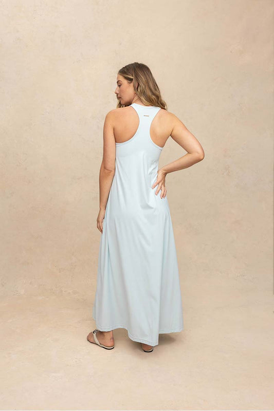 A woman modeling a light blue sleeveless maxi dress from the back, standing against a textured beige studio backdrop.