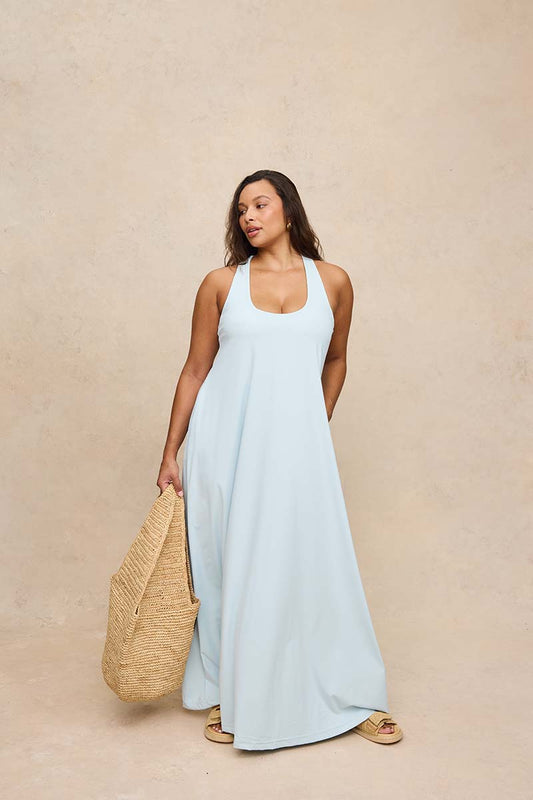A woman modeling a light blue halter-neck maxi dress, holding a large woven straw tote bag against a textured beige studio backdrop.