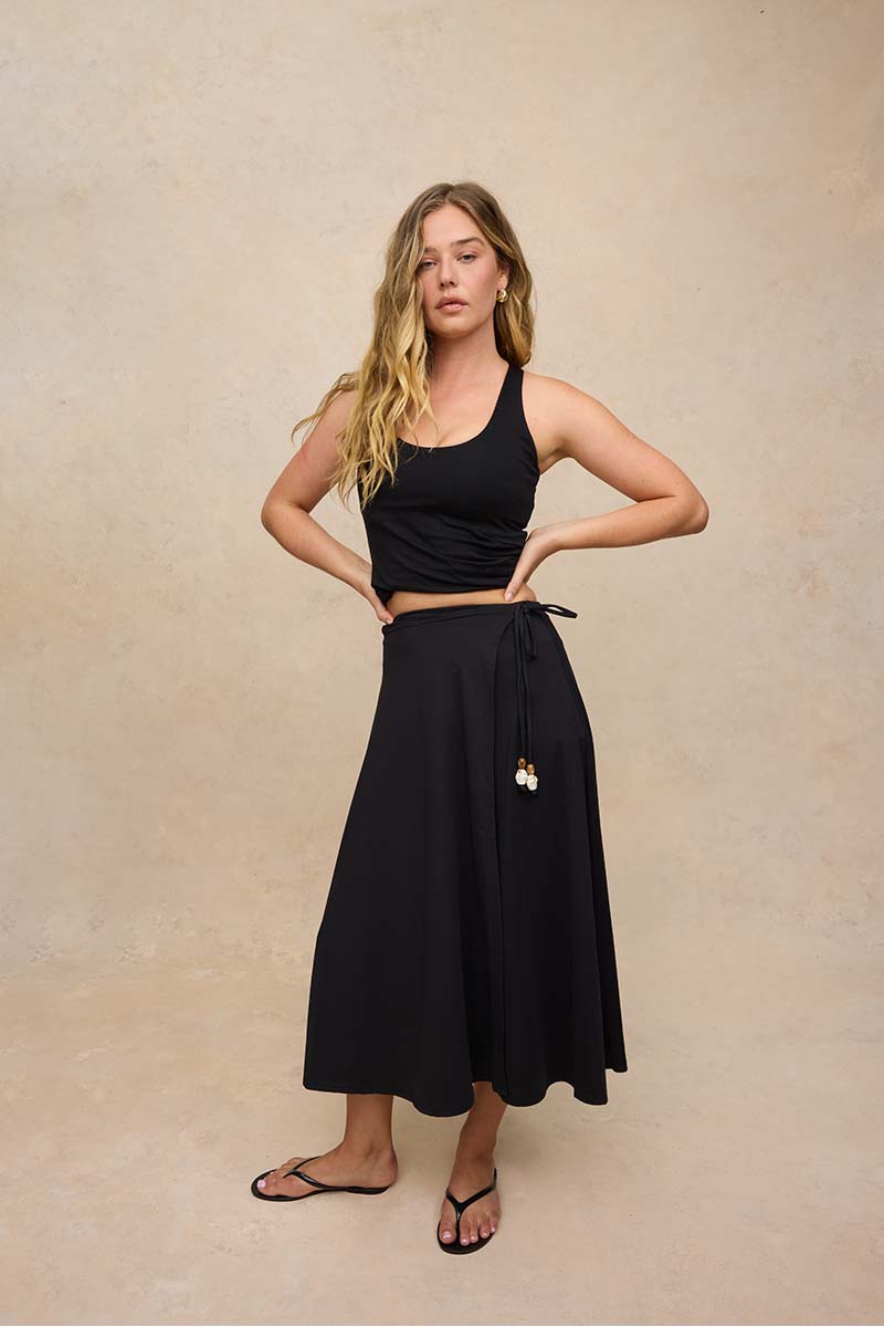 A woman modeling a black sleeveless crop top and a black high-waisted midi skirt with a drawstring and tassel detail, paired with black flip-flop sandals, against a textured beige studio backdrop.