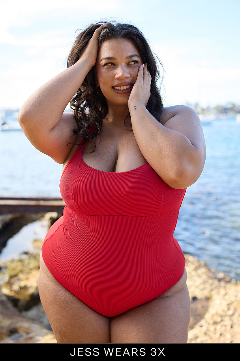 A woman in a red Adventure Suit by Fearless au stands smiling by the water, touching her face, with rocks and boats behind her. Text below reads: JESS WEARS 3X. Discover our limited edition swimwear collection.
