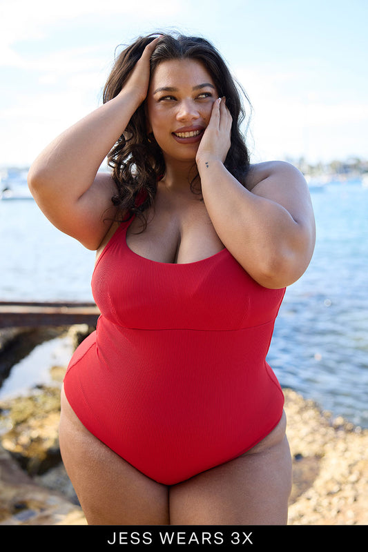 A woman in a red Adventure Suit by Fearless au stands smiling by the water, touching her face, with rocks and boats behind her. Text below reads: JESS WEARS 3X. Discover our limited edition swimwear collection.