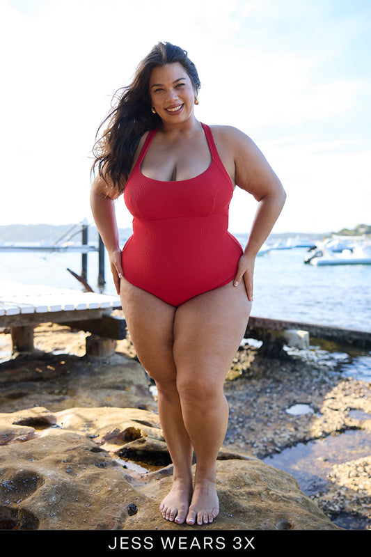 A smiling woman stands by the water, barefoot on rocks near boats and trees, wearing the bright red Adventure Suit by Fearless au. Text reads, JESS WEARS 3X.