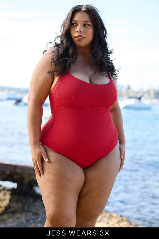 A woman with long dark hair stands by the water on a sunny day, wearing the limited edition Adventure Suit one-piece swimsuit from Fearless au. Text at the bottom reads, “JESS WEARS 3X.” Boats and trees are visible in the background.