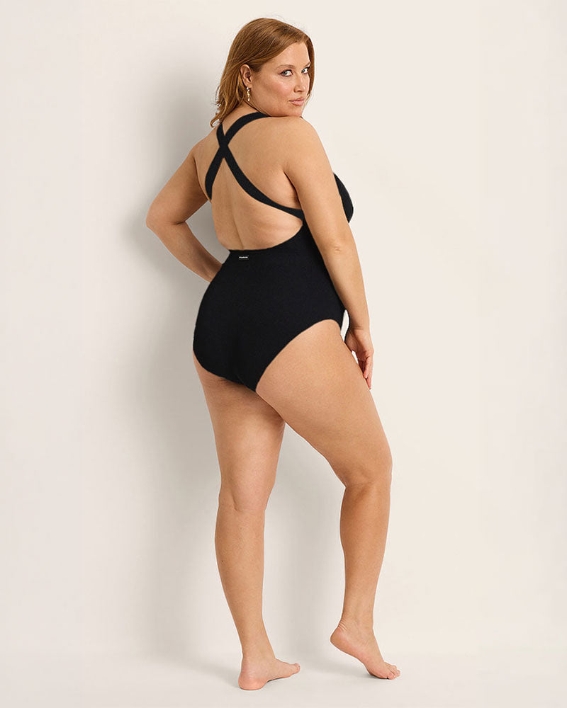 Woman wearing a supportive bust black one piece swimsuit against a plain background.