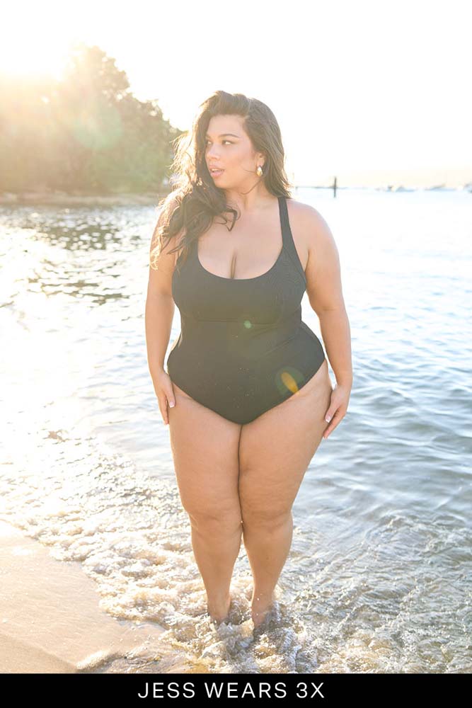 Woman in a black swimsuit standing by a body of water with a scenic background.