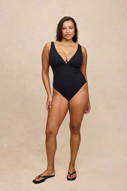A woman modeling a black one-piece swimsuit with a deep V-neckline and shoulder straps, standing against a neutral beige textured studio backdrop.