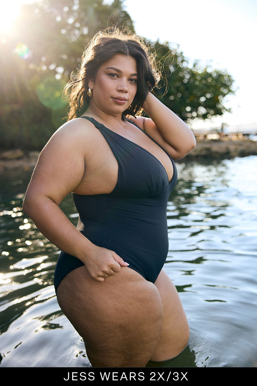 Woman in a navy swimsuit standing by water with trees in the background