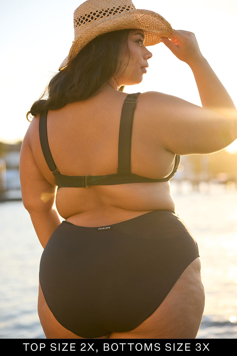 Woman wearing a black swimsuit and straw hat by a waterfront at sunset