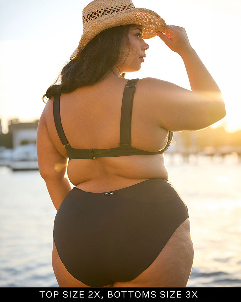 Woman in a Fearless black supportive bust and high waist one piece swimsuit standing by a waterfront, sunlit background