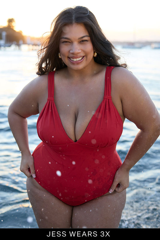 A woman smiles confidently in the Fearless au All Day One Piece—this supportive, tummy control swimsuit—while standing in shallow water at sunset. Text at the bottom reads, “JESS WEARS 3X.”.