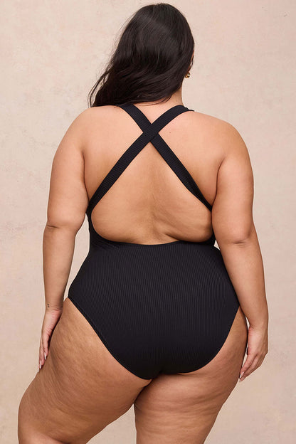 A plus-size model shown from the back wearing a black ribbed one-piece swimsuit with crisscross shoulder straps, set against a neutral beige textured background.