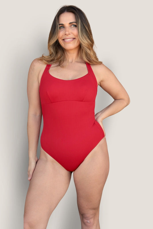 A woman with long, wavy hair smiles while wearing the red Fearless au Adventure Suit swimsuit, crafted from recycled ribbed fabric. She stands against a light background with one hand on her hip.