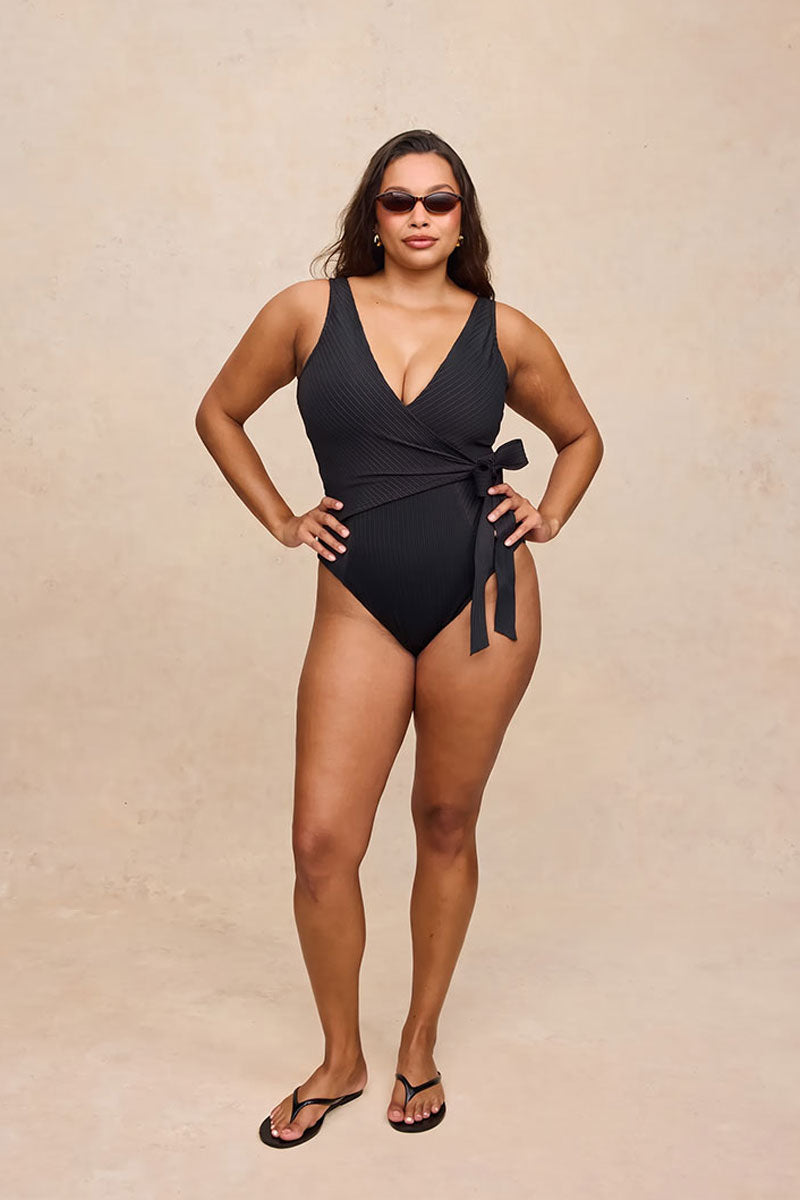 A woman modeling a black ribbed one-piece swimsuit with a wrap-style front and side tie, standing against a warm beige textured studio backdrop.