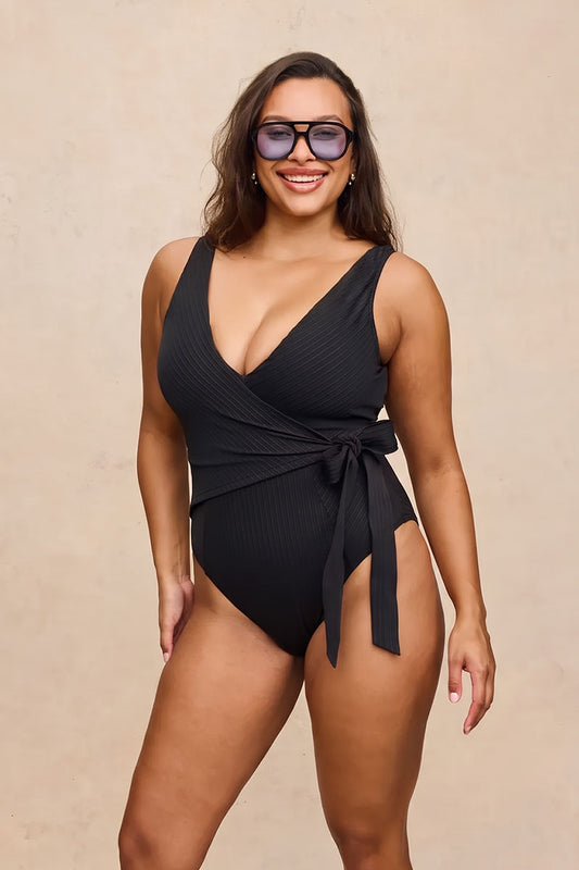A woman modeling a black ribbed one-piece swimsuit with a wrap-style V-neck bodice and side tie, photographed against a plain beige studio background.