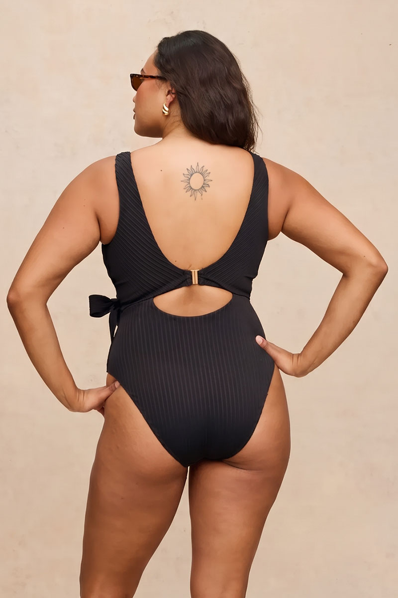 A woman modeling a black ribbed one-piece swimsuit from the back, featuring a low open back, side tie, and a small gold clasp, set against a warm beige background.