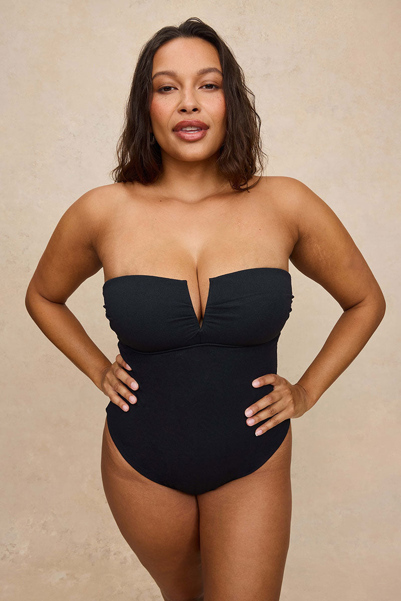 A woman modeling a strapless black one-piece swimsuit with a sweetheart neckline and a deep front keyhole cutout, posed against a textured beige studio backdrop.