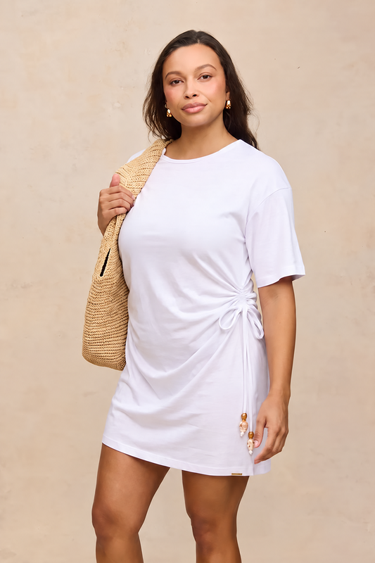 A woman modeling a white short-sleeve t-shirt dress with a side twist detail and small floral embroidery near the hem, carrying a woven straw tote bag against a textured beige wall.