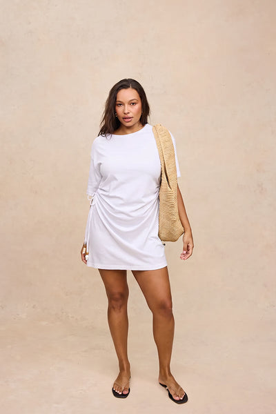 A woman modeling a white short-sleeve mini dress with side ruching, styled with a beige woven shoulder bag and black flip-flop sandals against a textured beige studio backdrop.