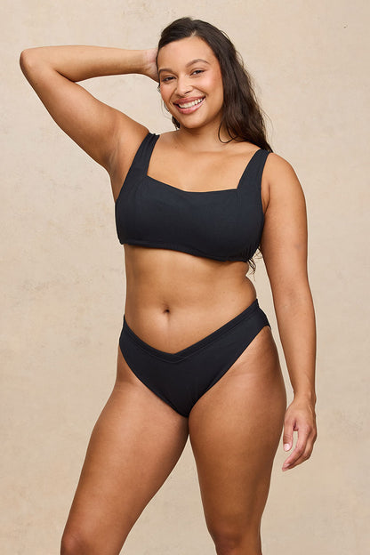 A woman modeling a black two-piece bikini set against a textured beige studio background. The set includes a scoop-neck bikini top with wide straps and matching high-cut bikini bottoms.