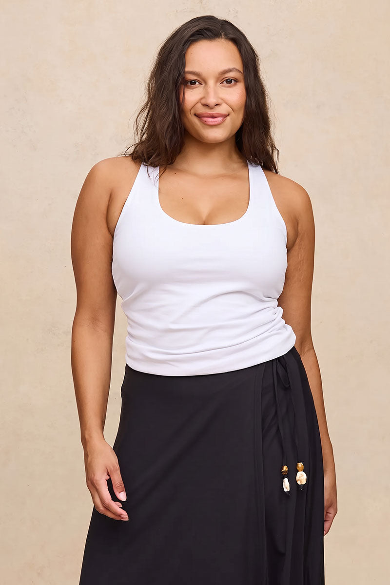 A woman modeling a white ribbed scoop-neck tank top and a black wrap skirt with tassel ties, set against a textured beige studio background.