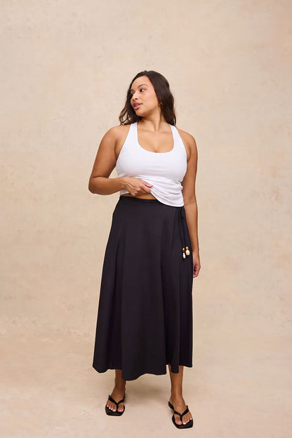 A woman modeling a black midi wrap skirt with a side tie and gold button accent, paired with a white tank top and black sandals, against a textured beige studio backdrop.