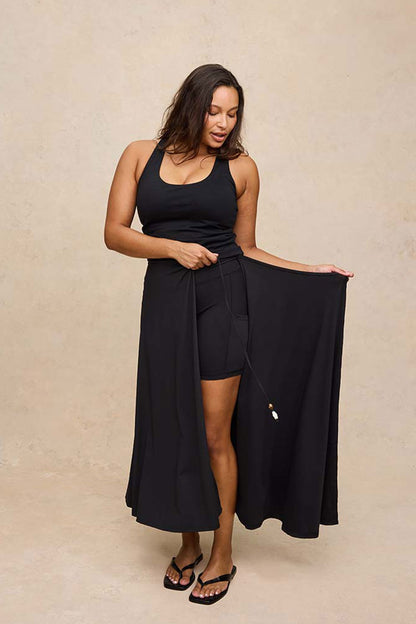 A woman modeling a black tank and skirt with a hidden shorts, holding out the side drape of the skirt against a textured beige studio backdrop.