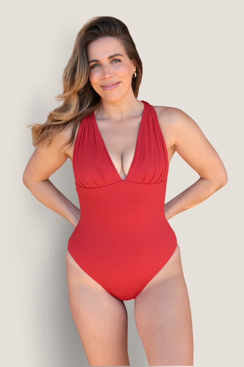 A woman with long, wavy brown hair smiles in a red Fearless au All Day One Piece - Standard swimsuit with built-in bust support and a deep V neckline, standing against a light background with her hands behind her back.