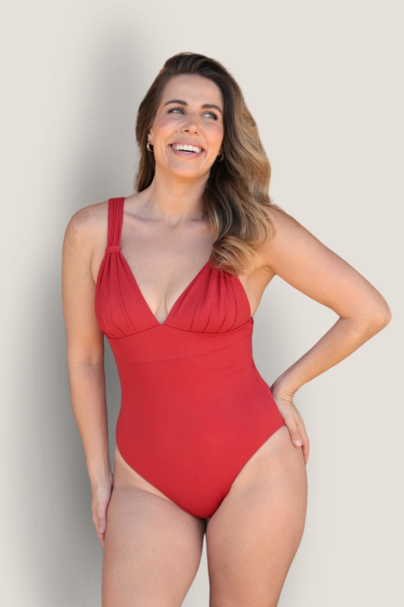 A woman with long wavy hair smiles while wearing the Fearless au All Day One Piece - Standard, a flattering swimsuit with tummy-hugging compression and thick straps, standing against a plain light gray background.