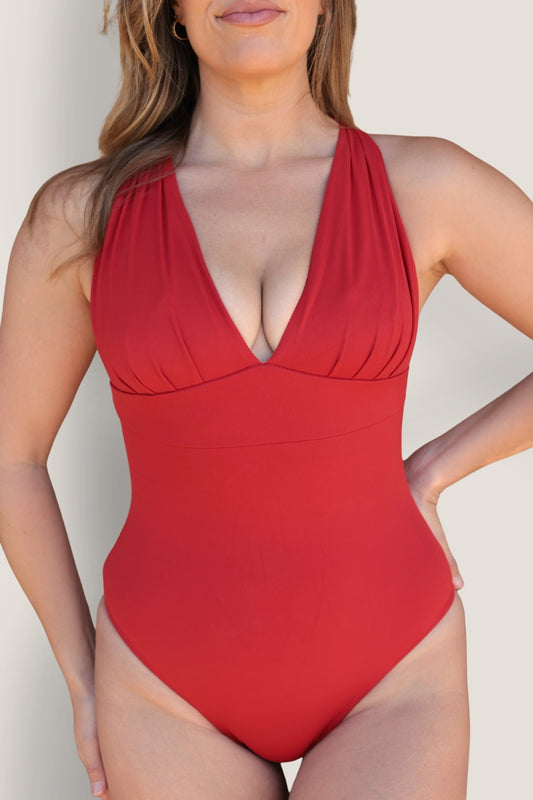 A woman models the Fearless au All Day One Piece - Standard, a red one-piece swimsuit with a deep V-neck, ruched details, and tummy-smoothing compression against a plain light background. Her face is partially visible.