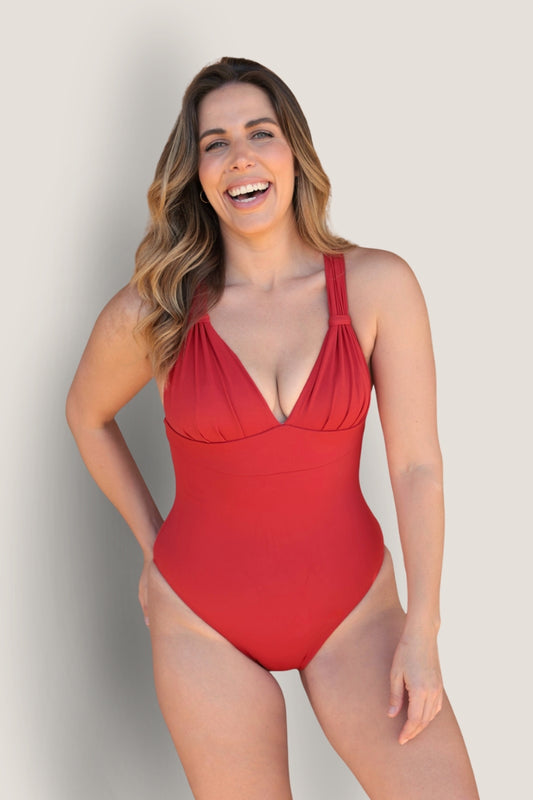 A smiling woman with long, wavy hair models the Fearless au All Day One Piece - Standard, a flattering red swimsuit with built-in bust support and wide shoulder straps, standing against a plain light background.