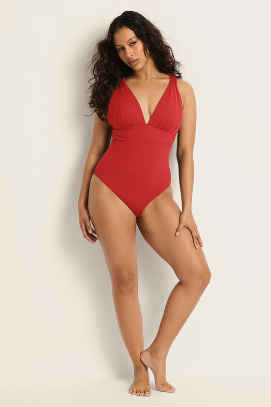 A woman with long, wavy hair smiles in the Fearless au All Day One Piece—a sleeveless, deep V-neck red swimsuit with tummy-hugging compression—posing against a plain, light background.