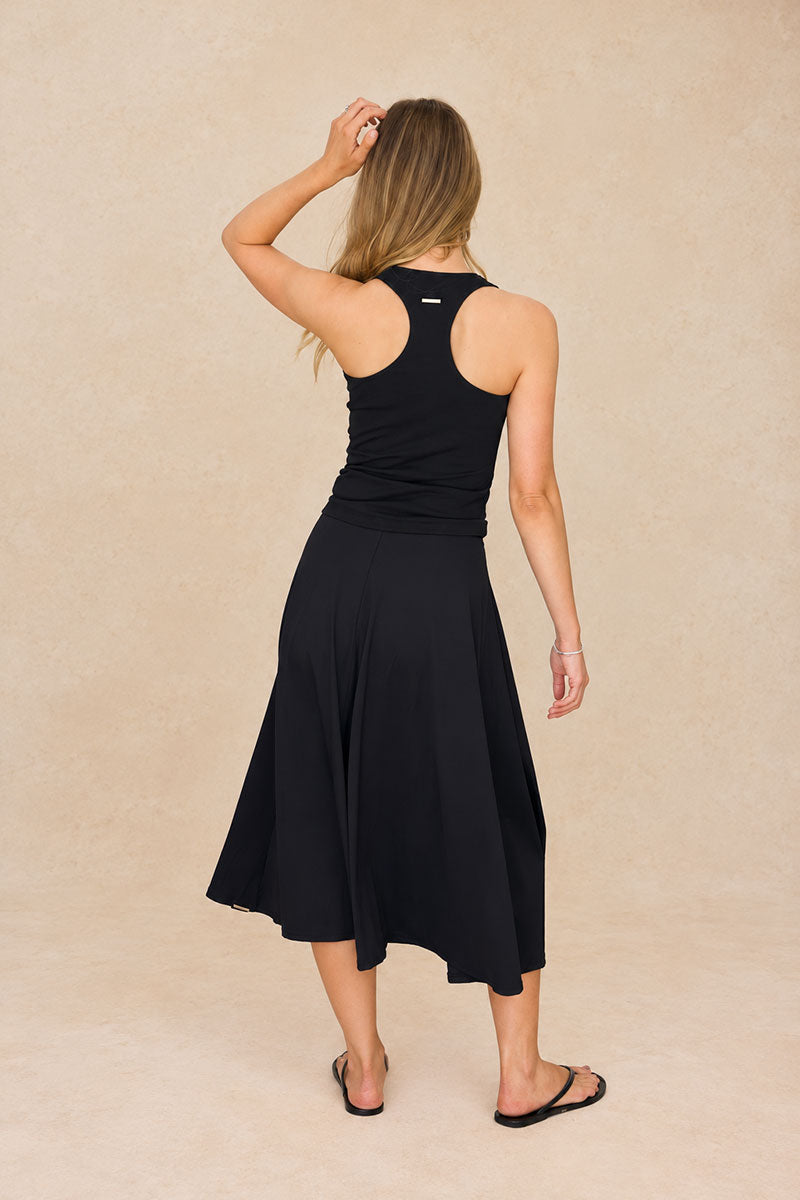 A woman is shown from the back wearing a black sleeveless midi dress with a racerback design and a flared skirt, paired with black flat sandals, against a textured beige studio backdrop.