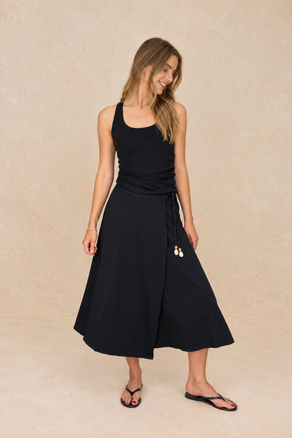 A woman modeling a black sleeveless midi wrap skirt with a tie waist and drawstring, paired with black flip-flops, against a textured beige studio backdrop.