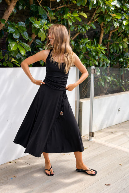 A woman modeling a black sleeveless midi dress with a gathered bodice and flowing skirt, standing outdoors on a light deck with green foliage and a white wall in the background.