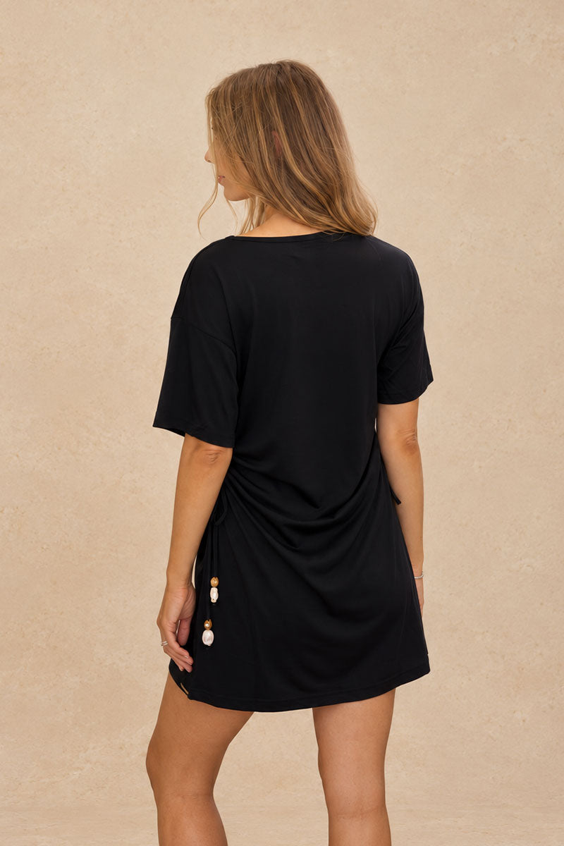 A woman shown from the back wearing a black short-sleeve mini dress with a gathered side detail and a decorative beaded tassel, set against a textured beige background.
