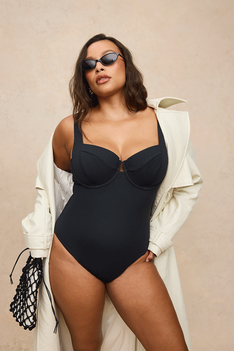 A woman modeling a black one-piece swimsuit with a keyhole cutout at the bust, styled with an oversized cream coat, sunglasses, and a black net bag against a beige textured background.