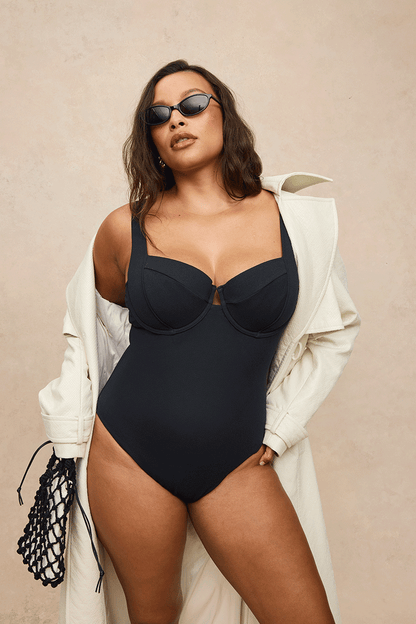 A woman modeling a black one-piece swimsuit with a keyhole cutout at the bust, styled with an oversized cream coat, sunglasses, and a black net bag against a beige textured background.