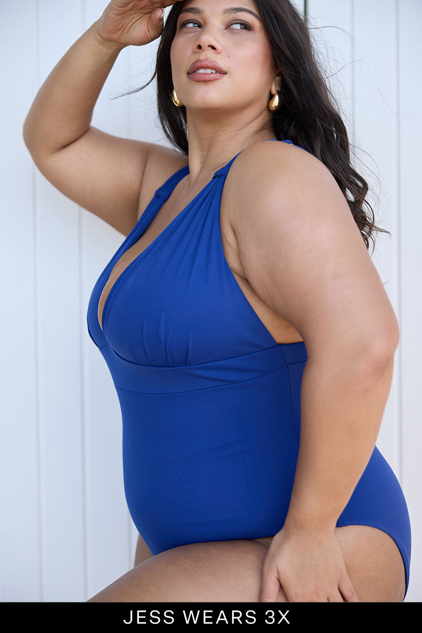 A woman with long, wavy dark hair and gold hoop earrings poses confidently in a bright blue Fearless au All Day One Piece swimsuit. Text below reads: JESS WEARS 3X.