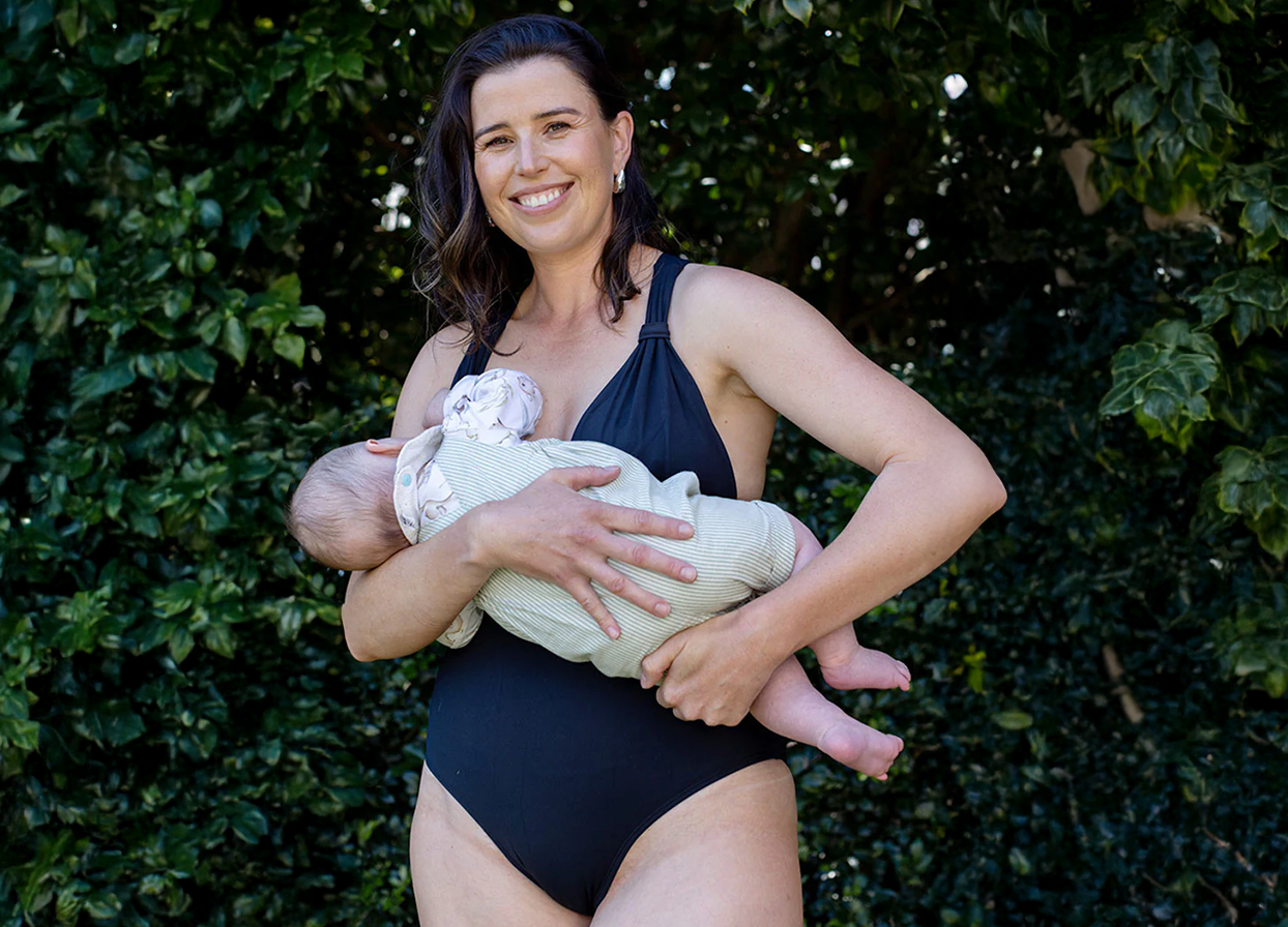 Woman in a black swimsuit holding a baby and breast feeding against a green leafy background

