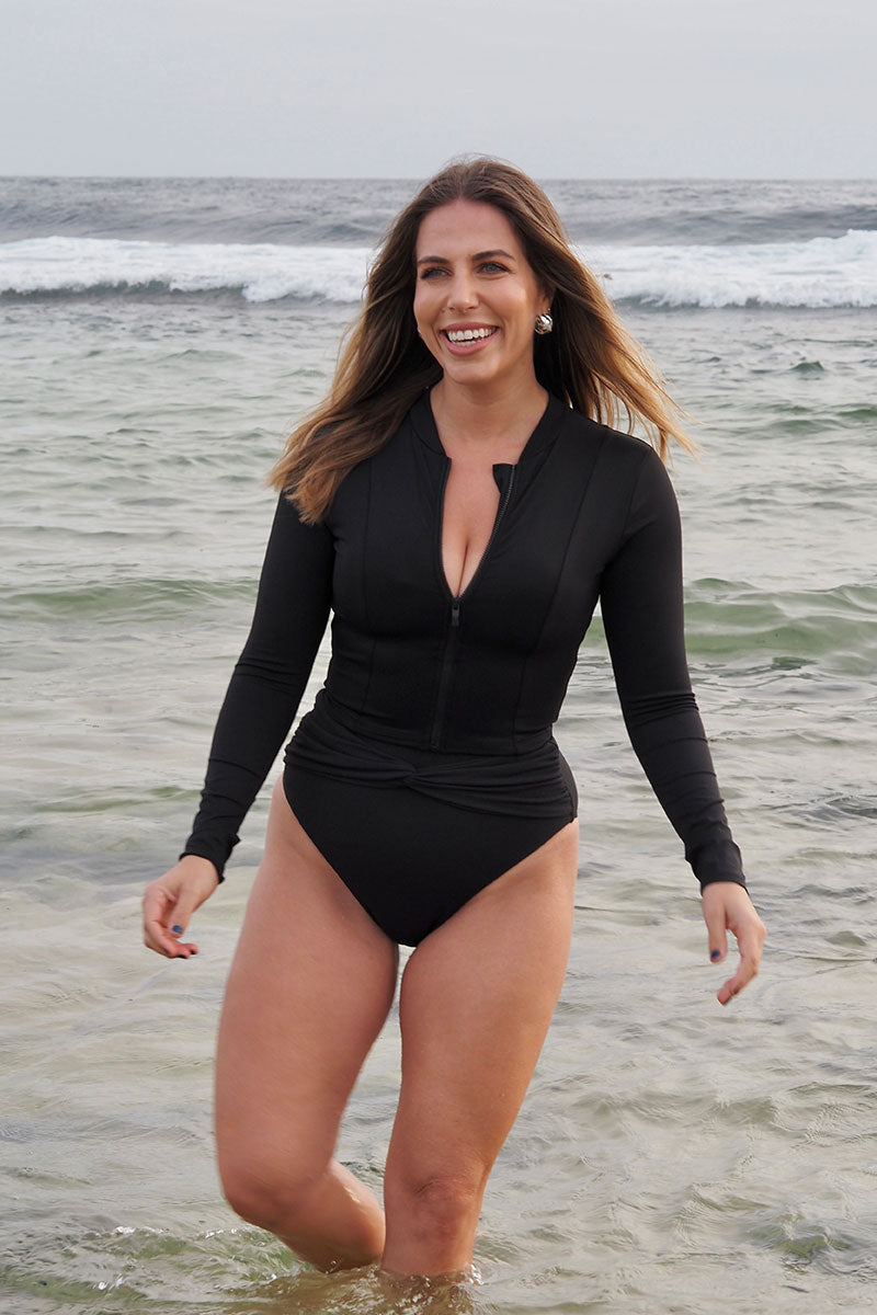 Blonde female standing in front of the ocean, wearing the Fearless black cropped rash vest with long sleeves and zip up front.