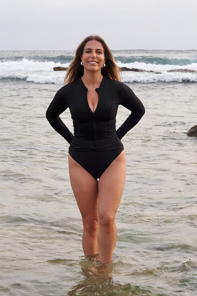 Blonde female standing in front of the ocean, wearing the Fearless black cropped rash vest with long sleeves and zip up front.