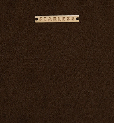 A close-up of a dark brown textured leather or faux-leather surface with a small rectangular metal plaque at the top center engraved with the word 'FEARLESS'.