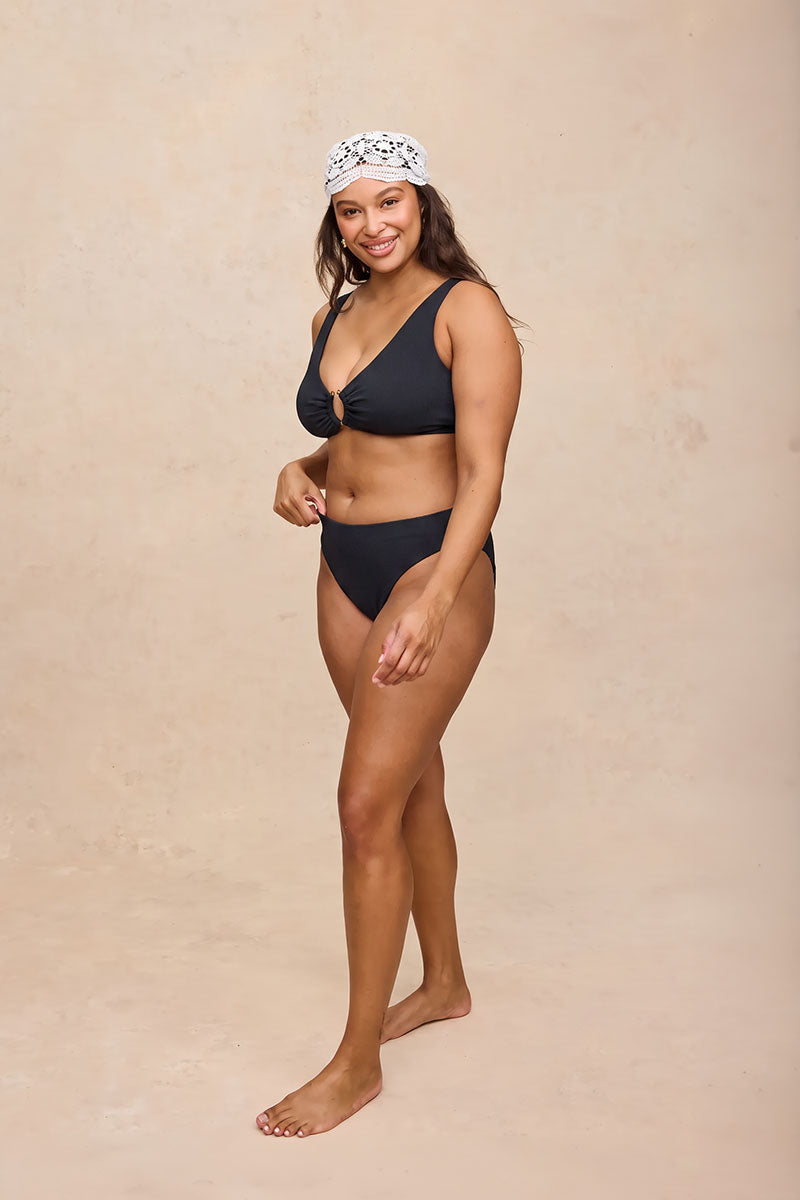 A woman modeling a two-piece black bikini with a matching black headband featuring a white pattern, standing against a textured beige studio backdrop.