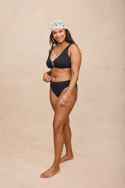 A woman modeling a two-piece black bikini with a matching black headband featuring a white pattern, standing against a textured beige studio backdrop.