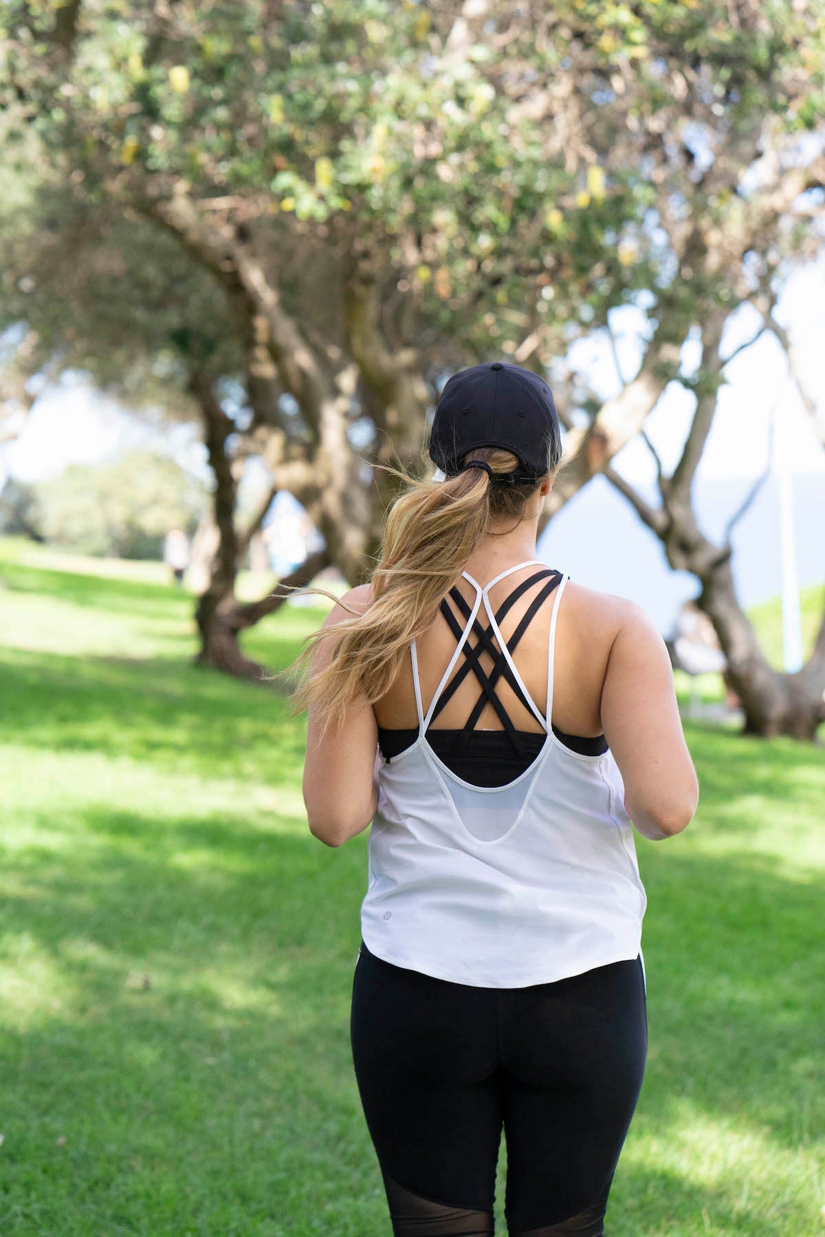 The back of a blonde haired woman wearing a black hat running. She is wearing a white singlet and black leggings. She is outdoors with trees in front of her.